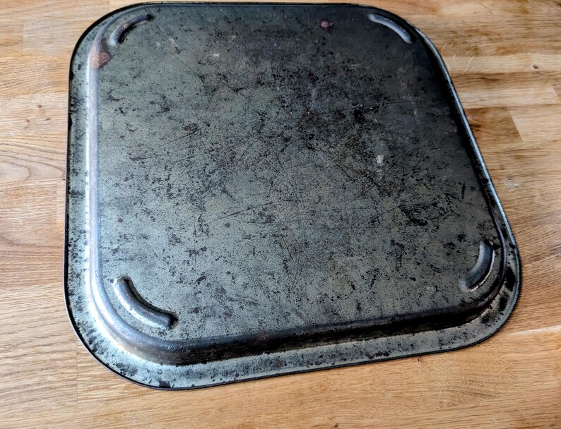 May include: A rectangular, vintage metal tray with rounded corners. The tray has a distressed, mottled gray finish with dark edges and visible scratches. The corners feature small, curved indentations.