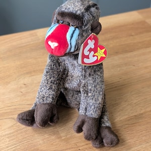 May include: A plush mandrill toy with mottled brown and gray fur, dark brown hands and feet, and a bright red nose with light blue accents. A heart-shaped red and yellow Ty Beanie Baby tag is attached to the ear of the stuffed animal.