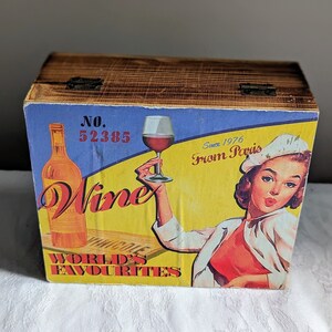 May include: A wooden box with a blue, yellow and red label. The label features a glass of red wine, a bottle of wine and a woman in a white hat. The text on the label reads "Wine World's Favourites" and "Since 1976 From Paris".