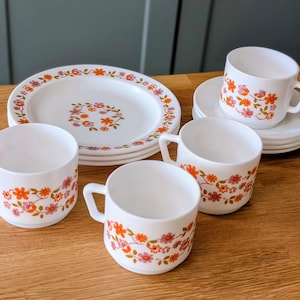 May include: A set of white ceramic tableware featuring a repeating floral pattern in orange, pink, and green. The collection includes three mugs with handles and a stack of matching plates and saucers, all decorated with small, delicate flower motifs.