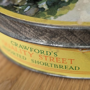 Puede incluir: Primer plano de una lata de galletas de mantequilla Crawford's Quality Street vintage. La lata tiene una etiqueta amarilla con texto verde y rojo. La tapa presenta una ilustración colorida. La lata es de metal y tiene un aspecto desgastado.