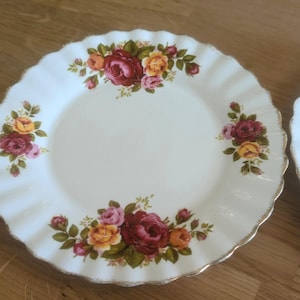 Può includere: Un piatto in porcellana bianca con bordo ondulato e un motivo floreale con rose rosse, gialle e rosa. Il piatto ha un bordo dorato.