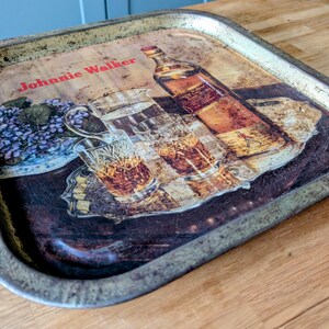 May include: Vintage metal serving tray with a printed design featuring a bottle of Johnnie Walker whiskey, glasses, and floral elements. The tray has a distressed, aged appearance with a metallic rim.