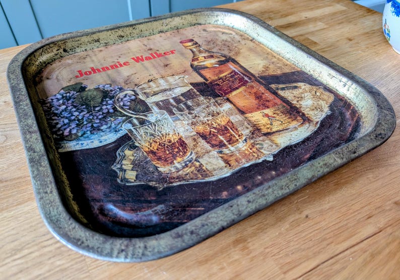 May include: Vintage metal serving tray with a printed design featuring a bottle of Johnnie Walker whiskey, glasses, and floral elements. The tray has a distressed, aged appearance with a metallic rim.