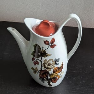 Puede incluir: Cafetera de cerámica blanca con un diseño floral con hojas marrones, naranjas y verdes y una flor blanca. La cafetera tiene una tapa roja con un pequeño agujero en la parte superior.