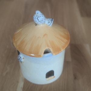 Può includere: Un zuccheriera in ceramica a forma di casetta per uccelli con un tetto marrone e un uccello bianco in cima. La casetta ha una piccola finestra e una base bianca.