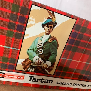 May include: A red and green tartan box featuring a man in a green kilt and a blue hat. The box reads "Crawford's Tartan Assorted Shortbread".