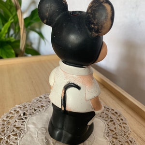 May include: A vintage Mickey Mouse figurine, featuring a black head with large ears, a white shirt, and black pants. The figurine has a curved black cane and is standing on a lace doily. The figurine is a collectible item.