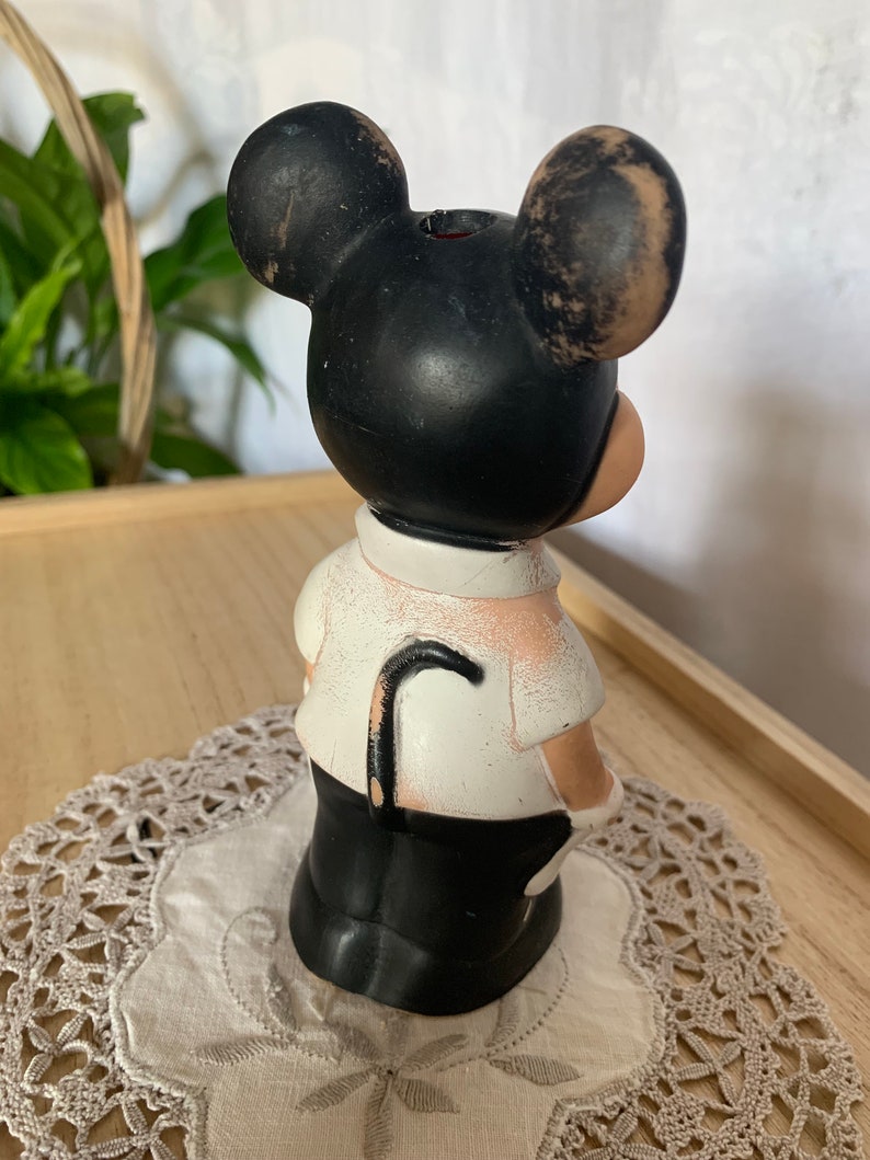 May include: A vintage Mickey Mouse figurine, featuring a black head with large ears, a white shirt, and black pants. The figurine has a curved black cane and is standing on a lace doily. The figurine is a collectible item.