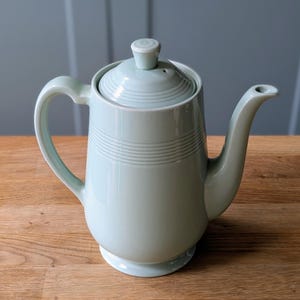 May include: A light mint green ceramic teapot featuring a cylindrical body with a series of horizontal embossed rings near the top, a curved handle, a matching lid with a small knob, and a tapered spout extending from the side of the vessel.