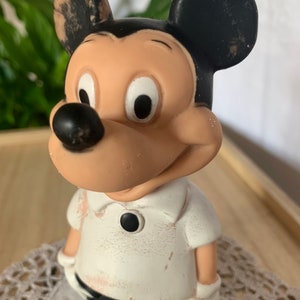 May include: A vintage Mickey Mouse figurine. The figure has a tan face, black ears, and a black nose. Mickey is wearing a white shirt with a black button and black pants. The figurine has white gloves and a smiling expression.