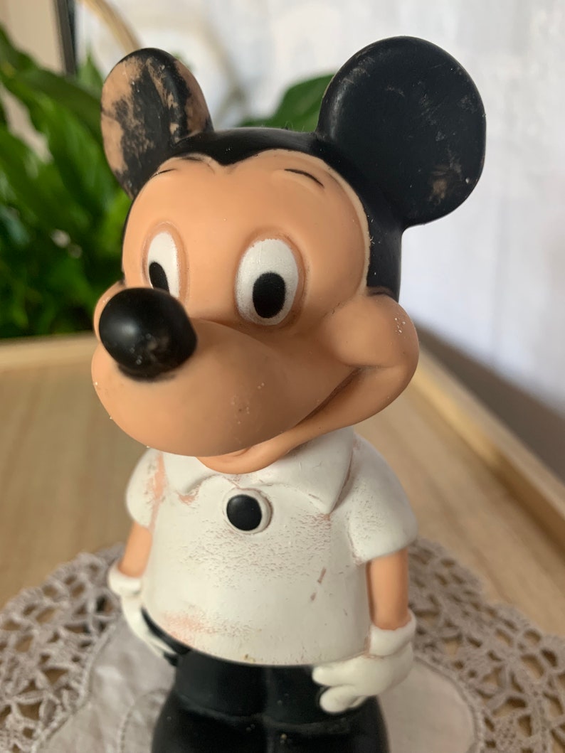 May include: A vintage Mickey Mouse figurine. The figure has a tan face, black ears, and a black nose. Mickey is wearing a white shirt with a black button and black pants. The figurine has white gloves and a smiling expression.