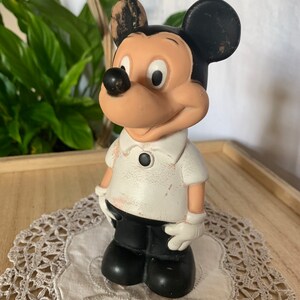 May include: A vintage Mickey Mouse figurine. The figure is wearing a white shirt with a black button, black shorts, and white gloves. The ears are black, and the face is tan with a black nose. The figurine is standing on a white doily.