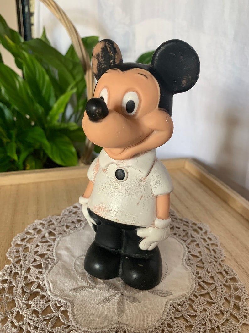 May include: A vintage Mickey Mouse figurine. The figure is wearing a white shirt with a black button, black shorts, and white gloves. The ears are black, and the face is tan with a black nose. The figurine is standing on a white doily.