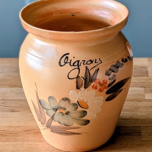 May include: A peach-coloured ceramic onion keeper with a wide opening and floral design. The word "Oignons" is written in black script on the side. The painted flowers include white, blue, and orange petals with green and brown leaves.
