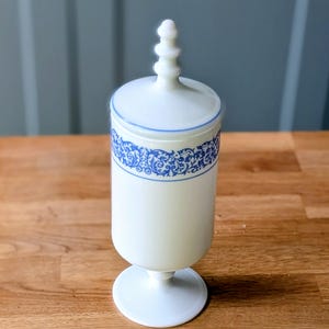 May include: A white, lidded apothecary jar with a blue floral design band. The jar has a pedestal base and a decorative finial on the lid. The design is a classic, elegant style.