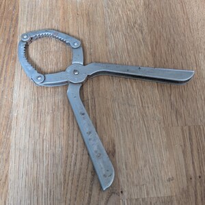 May include: A silver-coloured metal nutcracker featuring a circular, serrated gripping head and two long, flat handles connected by a central pivot point. The tool has a weathered, matte finish with visible signs of use and small surface imperfections.
