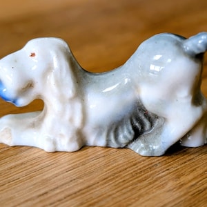 May include: A ceramic figurine of a dog in a reclining pose. The dog is primarily white with gray and blue accents. The figurine has a glossy finish and is resting on a wooden surface.