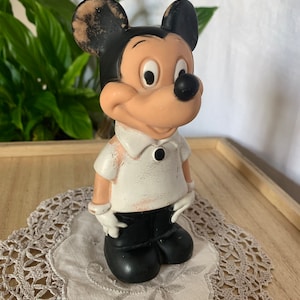 May include: Vintage Mickey Mouse figurine. The figure is painted with black, white, and tan colors. Mickey is wearing a white shirt, black pants, and white gloves. The figurine is standing on a lace doily.