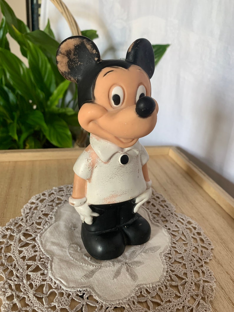 May include: Vintage Mickey Mouse figurine. The figure is painted with black, white, and tan colors. Mickey is wearing a white shirt, black pants, and white gloves. The figurine is standing on a lace doily.