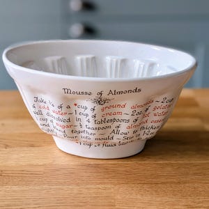 Puede incluir: Un molde de cerámica blanca con bordes acanalados que presenta una receta impresa para Mousse de Almendras en texto negro y rojo. La superficie exterior muestra instrucciones detalladas y medidas de ingredientes en unidades imperiales.