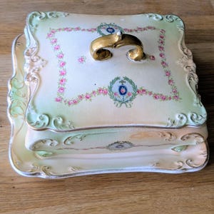 Pode incluir: Uma travessa quadrada vintage com tampa, com uma paleta de cores pastel creme, verde e rosa. A travessa é adornada com padrões florais e detalhes dourados, incluindo uma alça decorativa na tampa. As bordas têm um design ondulado.