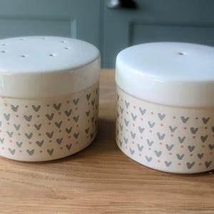 Op de afbeelding: Twee witte keramische containers met een grijs en roze hartpatroon. De containers hebben deksels met kleine gaatjes bovenop.