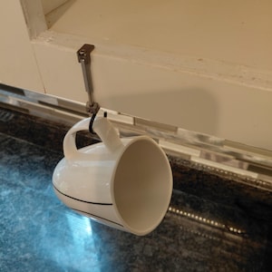 May include: A white ceramic mug with thin black horizontal lines hanging from a silver-toned metal hook. The hook is attached to the edge of a white cabinet frame, featuring a vertical support rod and a small screw mechanism for secure mounting.