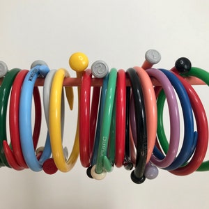 May include: A collection of colorful circular knitting needles with a hook on one end, hanging on a pink bar. The needles are made of plastic and come in a variety of colors, including green, red, blue, yellow, and pink.