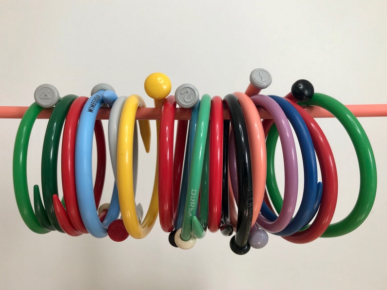 May include: A collection of colorful circular knitting needles with a hook on one end, hanging on a pink bar. The needles are made of plastic and come in a variety of colors, including green, red, blue, yellow, and pink.