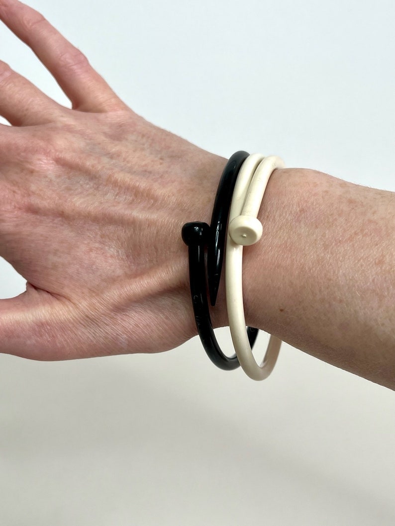 May include: Two bangle bracelets, one black and one white, with a nail-shaped design. The bracelets are worn on a person's wrist.