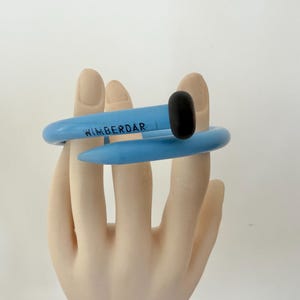 May include: A light blue nail-shaped bracelet featuring a smooth, curved design that wraps around the wrist. The head of the nail is black, and the word WIMBERDAR is printed in black capital letters along the side of the blue band.