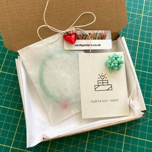 May include: A small cardboard box with a green and white tissue paper lining. The box contains a green bracelet and a card with a heart and the text "A gift for you - enjoy!"