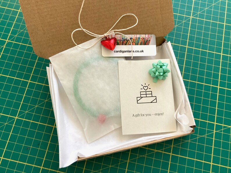 May include: A small cardboard box with a green and white tissue paper lining. The box contains a green bracelet and a card with a heart and the text "A gift for you - enjoy!"