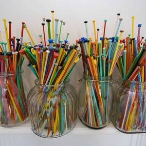 May include: Four glass jars filled with colorful knitting needles. The needles are arranged in a rainbow pattern and are all different lengths.