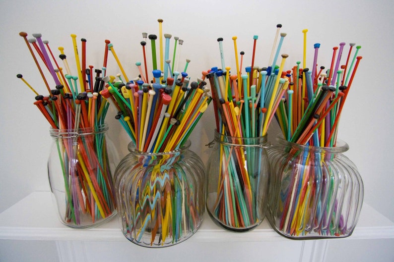 May include: Four glass jars filled with colorful knitting needles. The needles are arranged in a rainbow pattern and are all different lengths.