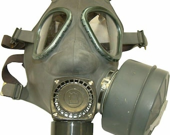 May include: A black gas mask with a large round filter attached to the side. The mask has two large eye pieces and a strap that goes around the head.