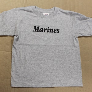 May include: A gray short-sleeved t-shirt with the word "Marines" printed in black on the front.