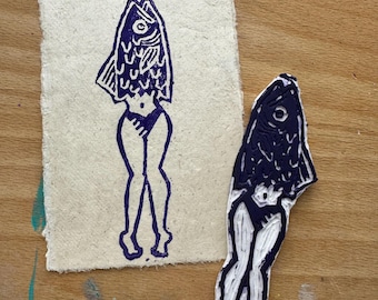 May include: A purple and white woodblock print of a person with a fish head and legs. The print is on a piece of white paper. The woodblock is also shown in the image.