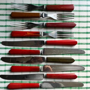 May include: A collection of vintage silverware, including forks and knives, arranged on a green and white checkered surface. The forks have tines and brown, red, and orange handles. The knives have stainless steel blades and matching handles.