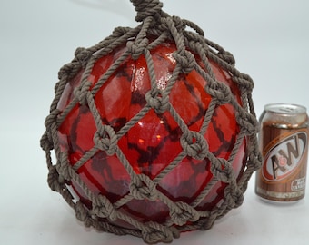 May include: A spherical red glass fishing float encased in a brown knotted rope net. The float features a dark, mottled pattern within the glass. An A&W root beer can is positioned to the right, providing a visual scale reference for the object.