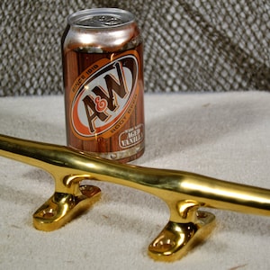 May include: A polished gold boat cleat with a rounded top and two mounting plates. The cleat is about 10 inches long and is sitting on a beige surface. A can of A&W Root Beer is in the background.