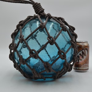 May include: A round, blue glass fishing float encased in a brown rope net. The float has a textured surface and is suspended by a matching rope. A brown and tan A&W soda can is next to the float.