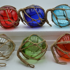 May include: Six glass fishing floats with a rope net. The floats are in various colors, including red, blue, green, and clear. The floats are all about the same size and have a rope handle.