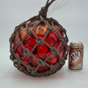 May include: A spherical red glass fishing float encased in a decorative brown knotted rope net. The float features a loop at the top for hanging. An A&W Root Beer soda can is positioned next to the float to provide a sense of scale for the object.
