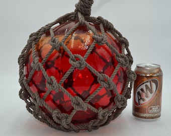 May include: A spherical red glass fishing float encased in a decorative brown knotted rope net. The float features a loop at the top for hanging. An A&W Root Beer soda can is positioned next to the float to provide a sense of scale for the object.