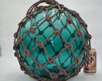 May include: A large, spherical, translucent teal glass float encased in a decorative brown knotted rope netting. The glass orb features a smooth, reflective surface and is positioned next to a standard 12-ounce A&W root beer soda can for scale.