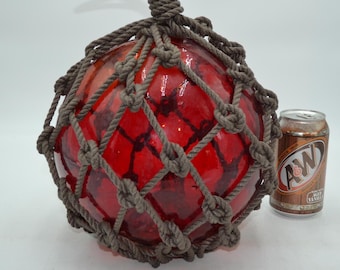 Può includere: Un grande galleggiante da pesca sferico in vetro rosso traslucido, racchiuso in una spessa rete di corda marrone annodata. L'oggetto è posizionato accanto a una lattina di alluminio da 355 ml di A&W Root Beer per un confronto dimensionale.