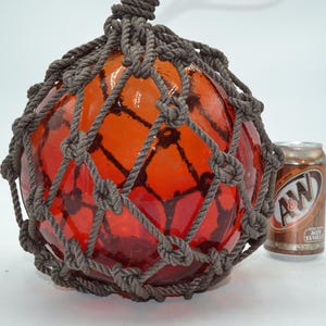 May include: A spherical orange glass fishing float encased in a dark gray knotted rope net. The float is positioned next to a standard-sized aluminum A&W Root Beer soda can for scale, providing a clear visual reference for its overall size.