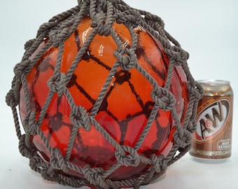 May include: A spherical orange glass fishing float encased in a dark grey knotted rope net. The float is positioned next to a standard-sized aluminium A&W Root Beer soda can for scale, providing a clear visual reference for its overall size.